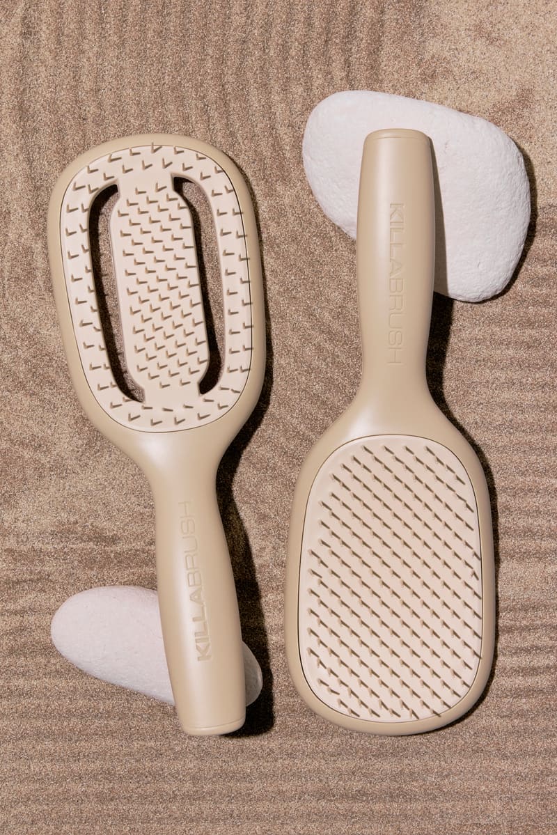 A cream KILLABRUSH vented brush, with an open centre, beside a cream paddle brush with a solid, cushioned head, on sand.